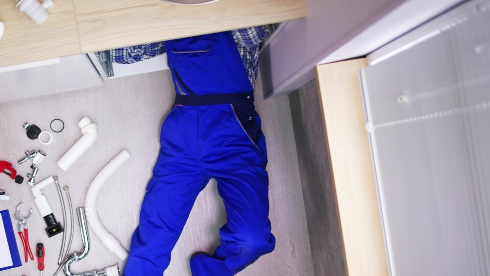 An ATC Plumbing plumber working under a commercial kitchen sink with tools and pipes scattered in LA County.