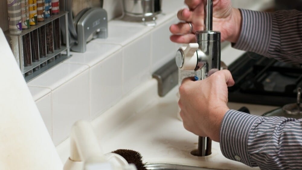 ATC Plumbing plumber working on a kitchen sink faucet, using a wrench to tighten or adjust the fixture in a modern kitchen setting.